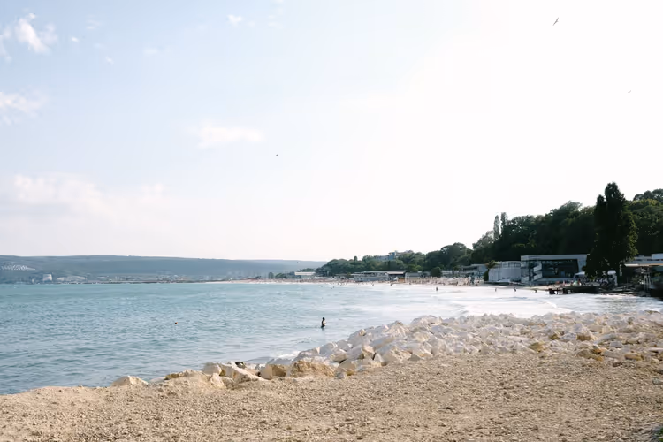 Sandstrand in Warna mit weißem Wellenbrecher aus Stein, ruhiger Schwarzmeerbucht und bewaldetem Ufer unter diesigem Himmel