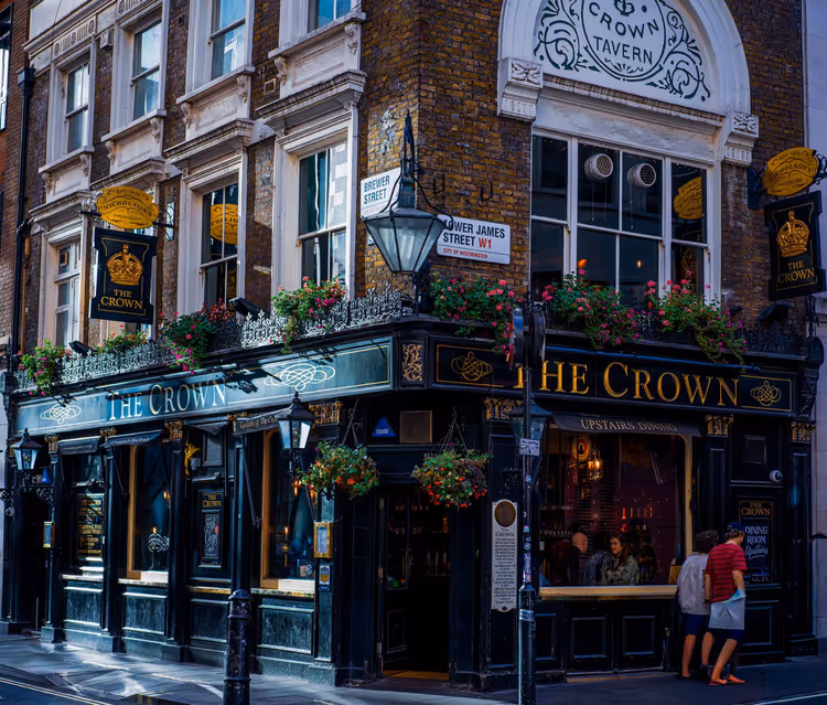 Schwarz-goldene Fassade des Pubs The Crown an einer Londoner Straßenecke mit Blumenampeln und Obergeschossen aus Backstein