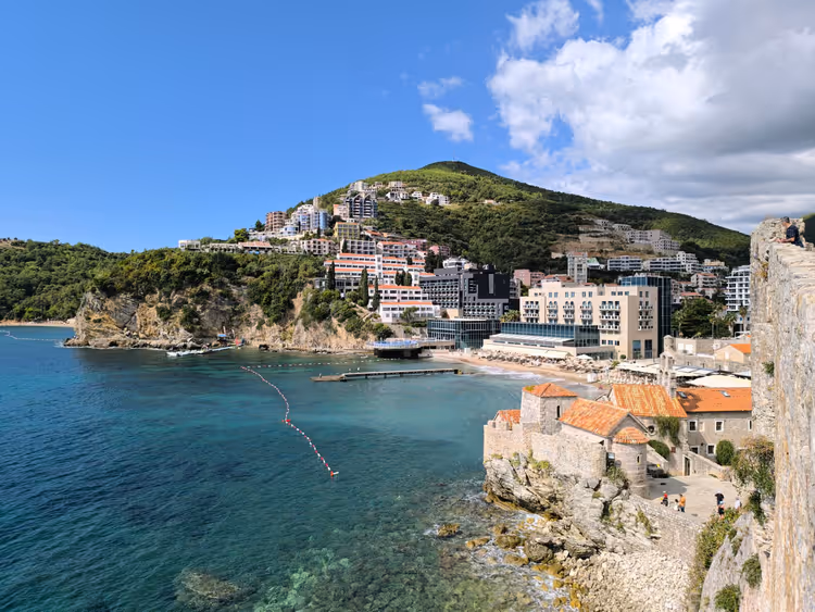 Baia turchese in Montenegro con hotel sul mare che risalgono una collina verde, vista da antiche mura di pietra