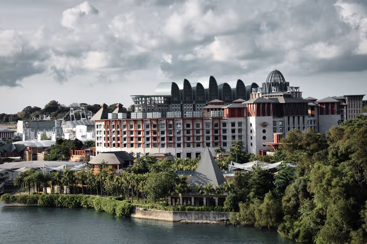Rot-weiße Hotelgebäude von Resorts World Sentosa über einer palmengesäumten Uferpromenade unter schweren Wolken in Singapur