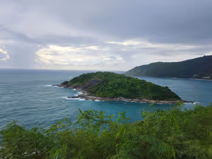 Kleine grüne Felseninsel vor der Küste von Phuket im türkisfarbenen Meer unter schweren grauen Wolken, Blick von einem begrünten Aussichtspunkt