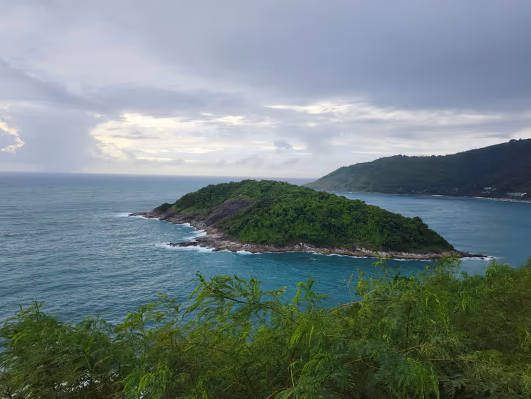 Kleine grüne Felseninsel vor der Küste von Phuket im türkisfarbenen Meer unter schweren grauen Wolken, Blick von einem begrünten Aussichtspunkt