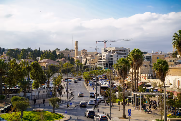 Straße in Israel mit Palmen, parkenden Autos und Steinhäusern, am Horizont ein Turm und ein Baukran