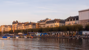 Seepromenade in Genf bei Sonnenuntergang mit vertäuten Booten, einer Reihe gestutzter Bäume und historischen Gebäuden im warmen Licht