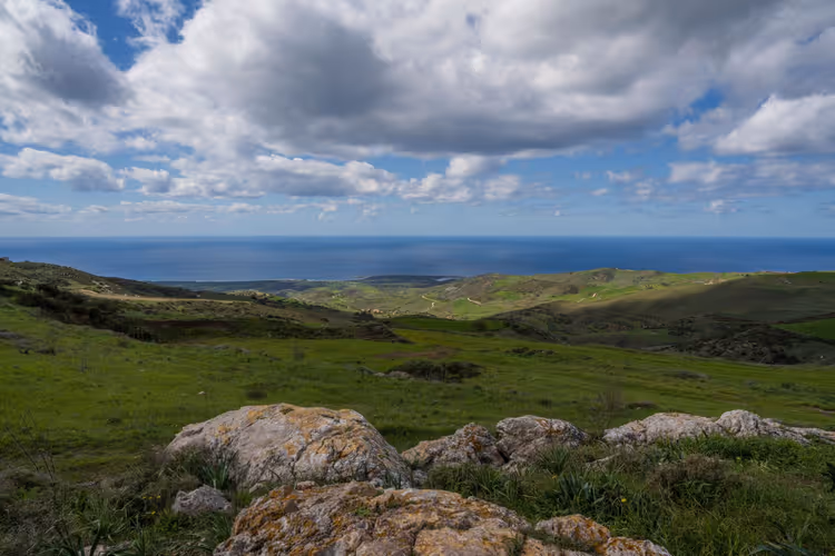 Grüne Hügel mit flechtenbewachsenen Felsen fallen unter schweren weißen Wolken zum Meer ab, Zypern