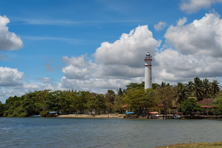 Weißer Leuchtturm über Palmen und grünen Bäumen am Ufer einer ruhigen Lagune mit kleinen Booten unter Haufenwolken