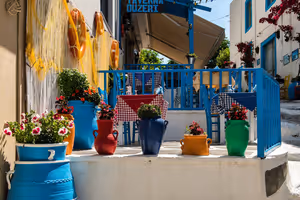 Terraza de una taberna griega con barandillas azules, manteles de cuadros y coloridas macetas de cerámica con flores en un escalón encalado