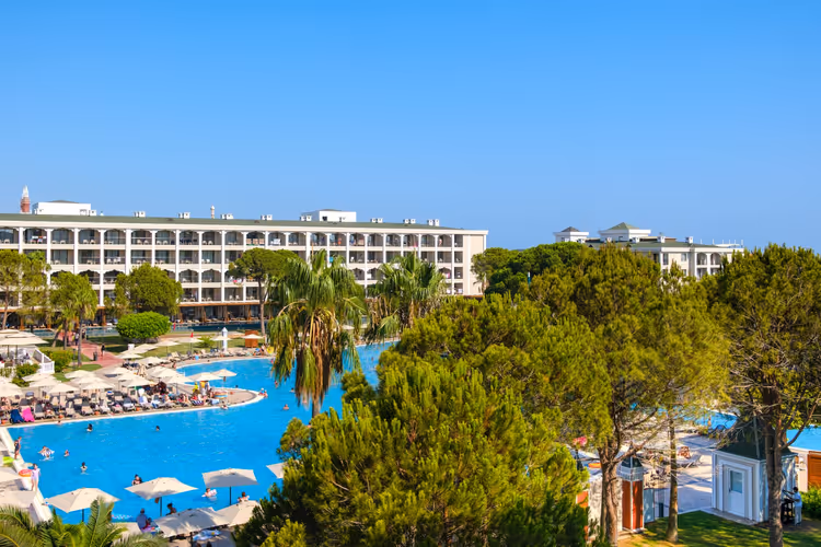 Großes Resorthotel mit weitem blauem Pool, Sonnenschirmen und Liegen zwischen Kiefern und Palmen in Belek