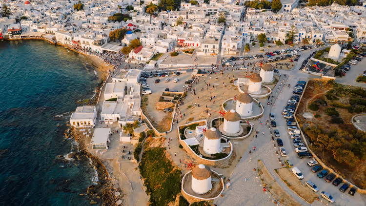 Luftaufnahme der Reihe runder, strohgedeckter Windmühlen über den weiß getünchten Häusern und der Uferpromenade von Mykonos-Stadt