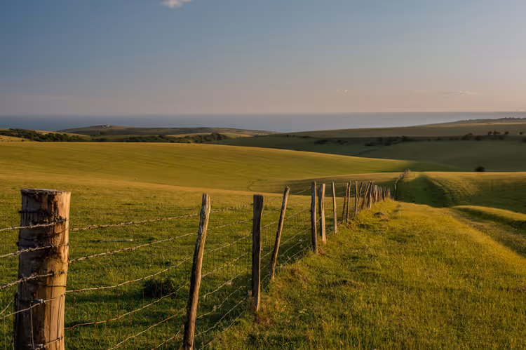Clôture en bois et fil barbelé longeant un sentier herbeux sur des collines vertes et vallonnées vers la mer, dans le sud de l'Angleterre