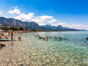 Badende im klaren, flachen Wasser vor einem Kiesstrand in Kemer, hinter der Küste schroffe Berge