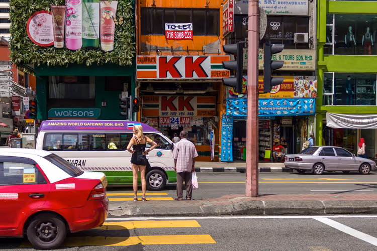 Due pedoni in attesa a un attraversamento di Kuala Lumpur davanti a vetrine colorate, un minibus e un taxi rosso