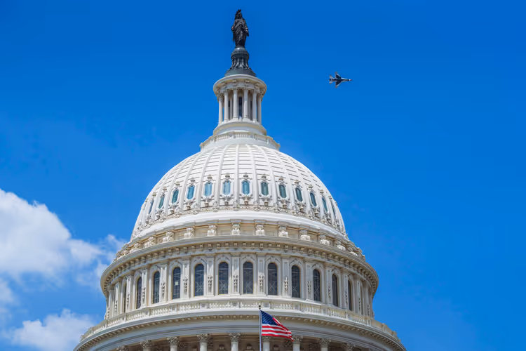 Weiße Kuppel des Kapitols in Washington vor tiefblauem Himmel, darunter eine US-Flagge, ein Düsenflugzeug fliegt vorbei
