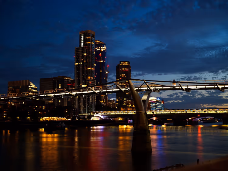 Millennium Bridge über der Themse bei Nacht in London, beleuchtete Hochhäuser spiegeln sich im dunklen Wasser