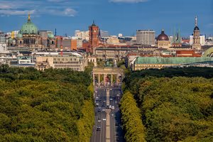 Berliner Skyline über den Baumkronen des Tiergartens, mit Brandenburger Tor am Ende einer breiten Allee und Domkuppel dahinter