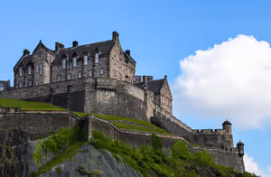 Castello di Edimburgo con mura di pietra e torrette su una rupe rocciosa ed erbosa contro un cielo azzurro con una nuvola bianca