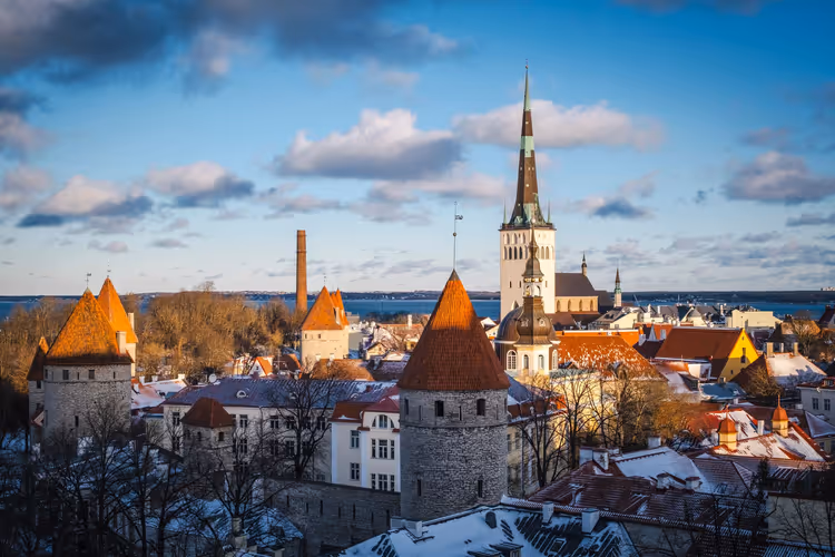 Mittelalterliche Türme mit roten Kegeldächern und hoher Kirchturm in der Altstadt von Tallinn, Schnee auf Dächern, dahinter Meer