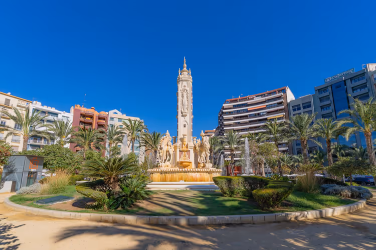 Reich verzierter Steinbrunnen mit hohem Mittelturm, umgeben von Palmen und Wohnblocks in Alicante