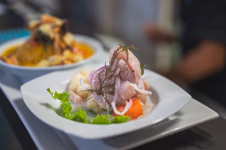 Peruanisches Ceviche mit rohem Fisch, roten Zwiebeln, Süßkartoffel und Salat auf weißem Teller, dahinter unscharf ein weiteres Gericht