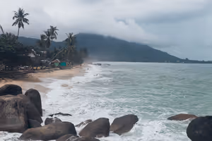 Wellen brechen über dunkle Felsblöcke an einem palmengesäumten Strand auf Koh Samui, tiefe Wolken verhüllen einen grünen Hügel