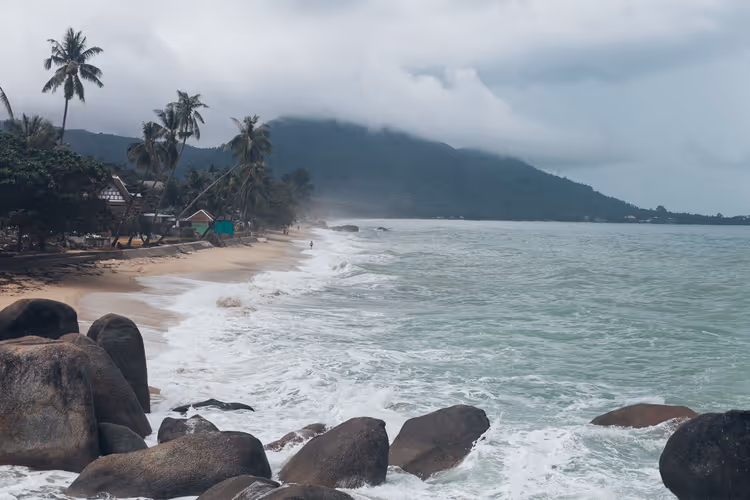 Wellen brechen über dunkle Felsblöcke an einem palmengesäumten Strand auf Koh Samui, tiefe Wolken verhüllen einen grünen Hügel