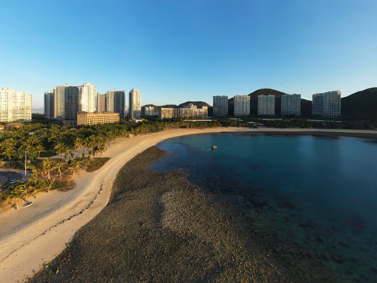 Luftaufnahme einer geschwungenen Sandbucht auf Hainan, gesäumt von Palmen und Resort-Hochhäusern unter klarem Himmel