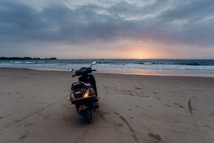 Motorroller auf einem breiten Sandstrand in Goa, zum Meer hin geparkt, bei Sonnenuntergang unter schweren Wolken