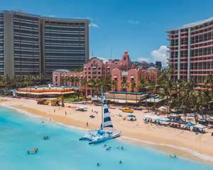 Rosa Hotel mit Glockenturm zwischen Palmen an einem Sandstrand, am türkisfarbenen Ufer ein Katamaran mit gestreiftem Segel