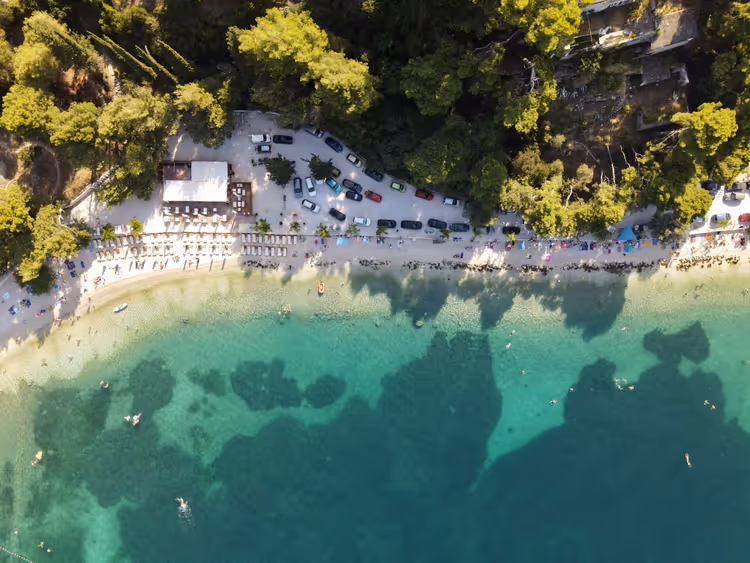 Flyfoto av en rullesteinsstrand med parasoller, sjø og parkering ved furutrærne