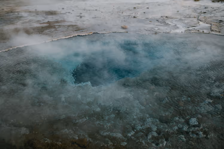 Dampf steigt aus einer türkisfarbenen heißen Quelle mit mineralverkrustetem Rand in einem isländischen Geothermalgebiet