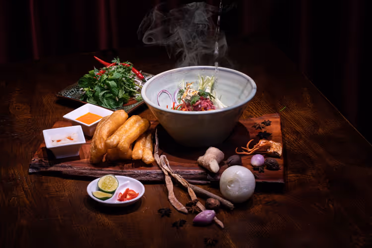 Dampfende Schale vietnamesischer Nudelsuppe auf einem dunklen Holztisch mit frittierten Teigstangen, Kräutern, Limette und Gewürzen
