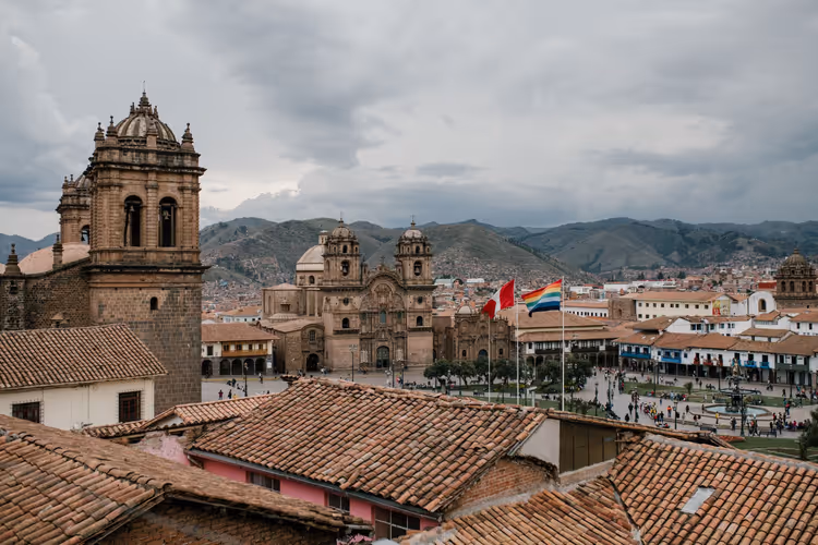 Terrakottadächer und steinerne Kirchtürme am Hauptplatz von Cusco mit Fahnen und Bergen unter grauen Wolken