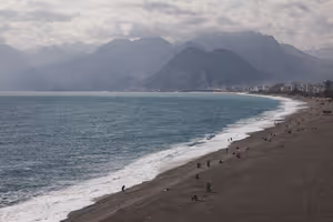 Langer dunkler Kieselstrand mit Brandung und vereinzelten Menschen unter dunstigen Bergen und schweren Wolken an der türkischen Küste