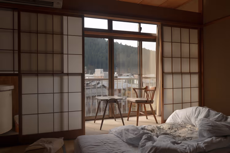 Schummriges traditionelles japanisches Zimmer mit Shoji-Schiebewänden, zerwühltem Futon sowie Holzstuhl und Tisch am Fenster, Japan