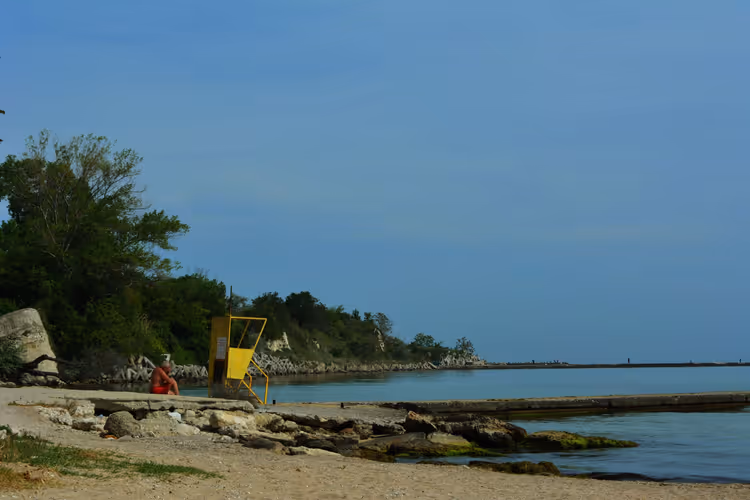 Ruhiger Sandstrand in Bulgarien mit gelbem Rettungsturm, Felsen, Betonmole und bewaldeter Landzunge am Schwarzen Meer