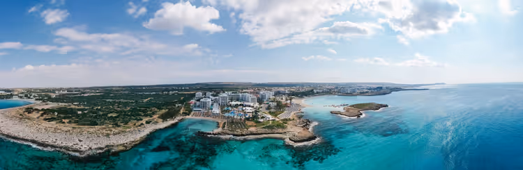 Luftpanorama einer Ferienküste auf Zypern mit Hotels, felsigem Ufer, einer kleinen Insel und türkisfarbenem Meer