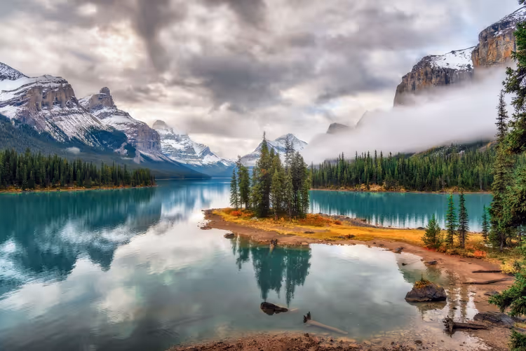 Spirit Island mit Fichtengruppe im türkisfarbenen Maligne Lake, dahinter leicht verschneite Gipfel und tiefe Wolken, Jasper