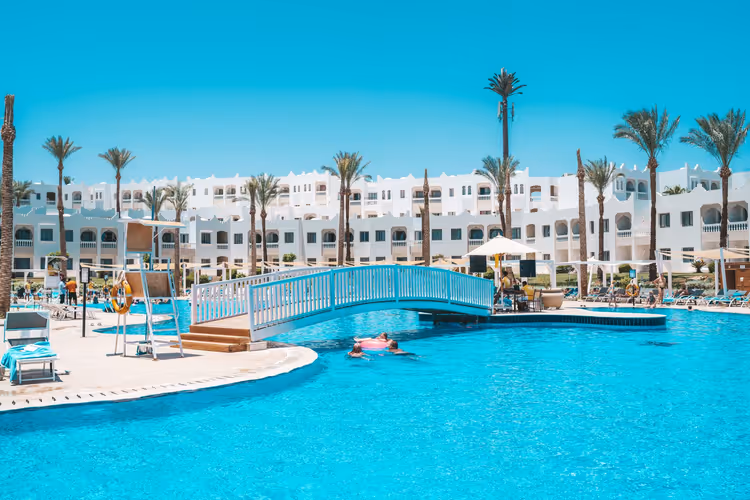 Türkisfarbener Hotelpool mit weißer Fußgängerbrücke, Palmen und niedrigen weißen Gebäuden in Scharm El-Scheich
