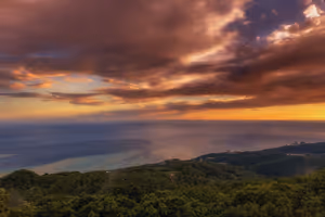 Sonnenuntergang über dem Meer, von bewaldeten Hügeln gesehen, mit schweren orangefarbenen und violetten Wolken über dunstiger Küste