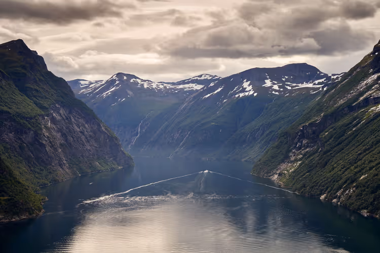 Norwegischer Fjord zwischen steilen grünen Felswänden mit der Kielspur eines Bootes auf dem Wasser, dahinter Gipfel mit Schneeresten