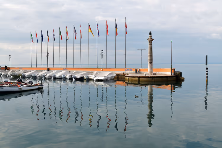 Reihe von Nationalflaggen und abgedeckten Booten an einem Hafensteg, gespiegelt im ruhigen Wasser des Gardasees