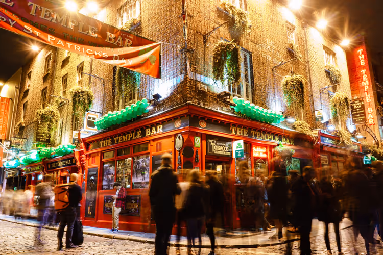 Rote Fassade des Temple Bar Pubs in Dublin bei Nacht, geschmückt mit grünen Ballons und Lichtern, davor verschwommene Passanten